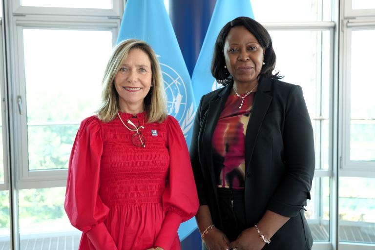 Two women standing side by side in front of large windows and blue United Nations flags, posing for a formal photograph.