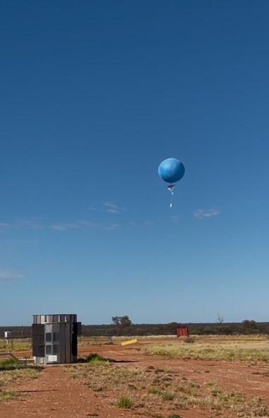 A blue weather balloon floats in the clear sky above a flat, grassy landscape with scattered equipment on the ground.