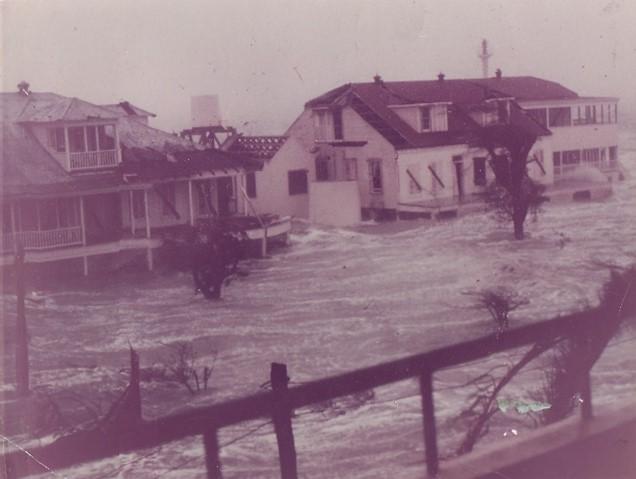 Floodwaters partially submerge houses, with strong currents and visible damage to buildings and trees, under overcast skies.