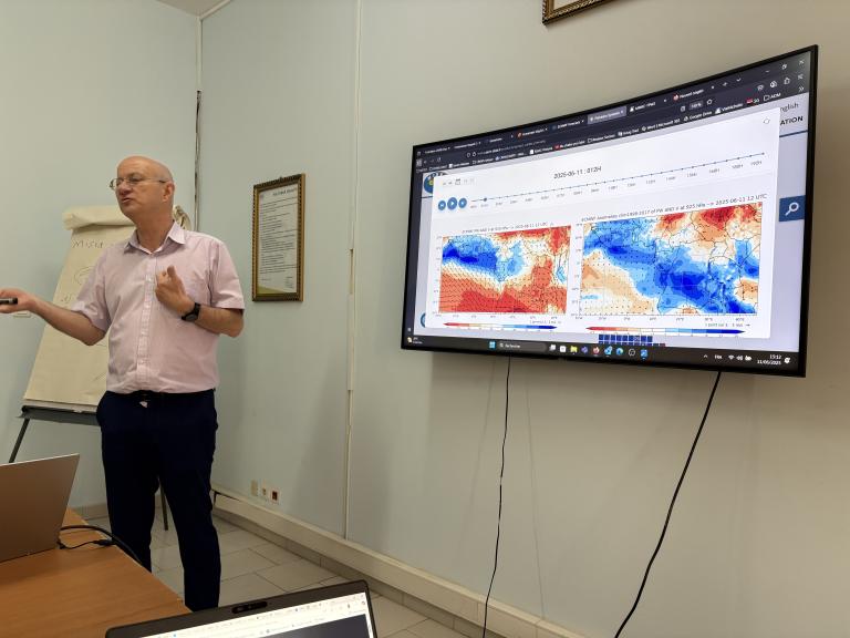 A man gives a presentation in a room, standing next to a screen displaying weather maps with red and blue color coding.