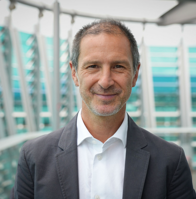 A middle-aged man with short dark hair and a beard smiles at the camera, wearing a white shirt and gray blazer, standing in front of a modern glass building.