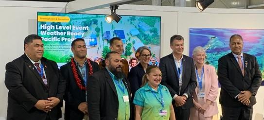 A group of people in business attire and uniforms pose for a photo in front of a display reading "High Level Event Weather Ready Pacific Programme".
