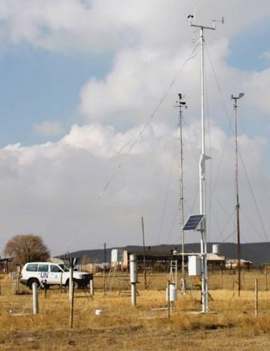 A weather monitoring station with multiple sensors and a solar panel stands in a dry field; a white UN vehicle is parked nearby.