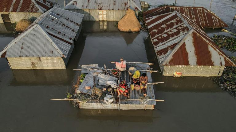 A group of people sits on a makeshift raft with belongings, surrounded by flooded tin-roof houses in a waterlogged area.