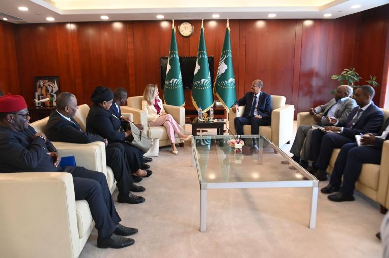 A group of officials sit in a formal meeting room, engaged in discussion around a glass coffee table, with flags and a clock displayed in the background.