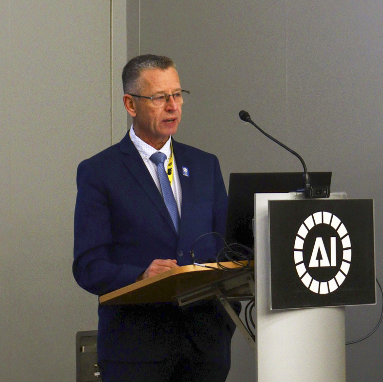 A man in a suit stands at a podium with a microphone, speaking in front of a sign featuring the letters "AI.