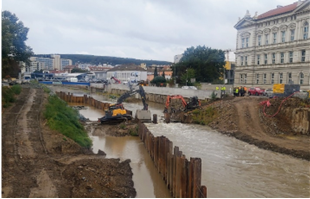 Excavators and workers are reinforcing a riverbank with steel sheet piles at a construction site alongside a city street and large building.
