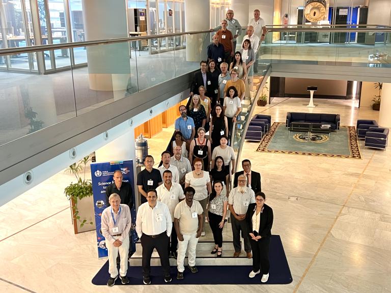 A group of people pose for a photo on a staircase and landing in a modern building, with conference badges visible and a WHO Science Hub banner nearby.