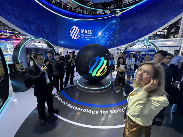 People gather around a digital display featuring a large black sphere with the "MAZU" logo at a technology expo, with informational screens and blue lighting in the background.