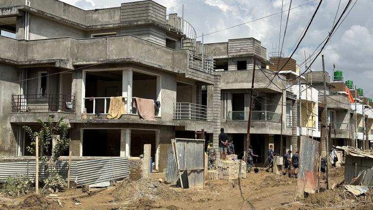 Row of unfinished concrete houses under construction, with construction workers at the site and laundry hanging from a balcony, in a muddy, cluttered area with exposed wiring and debris.