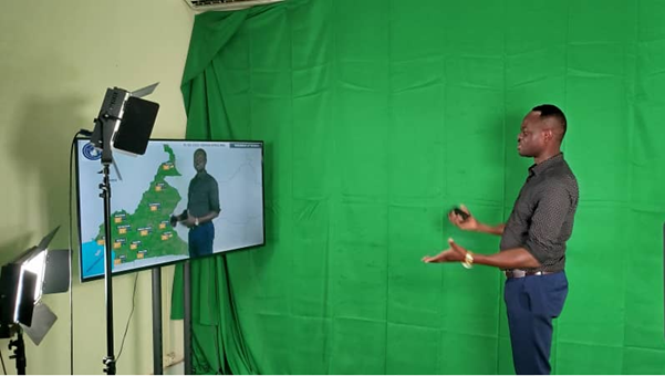 A man stands in front of a green screen presenting a weather forecast, with studio lights and a monitor displaying a map beside him.