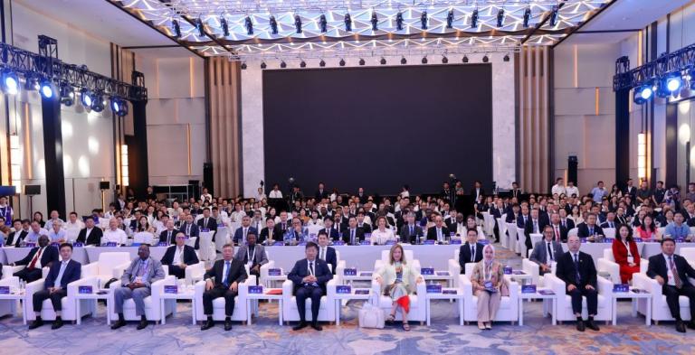 A large group of people, including officials seated in front rows, attend a formal conference in a modern, well-lit auditorium with a blank screen on stage.