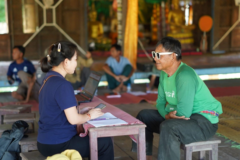 A woman interviews a man in sunglasses at a table indoors; other people sit in the background on mats.