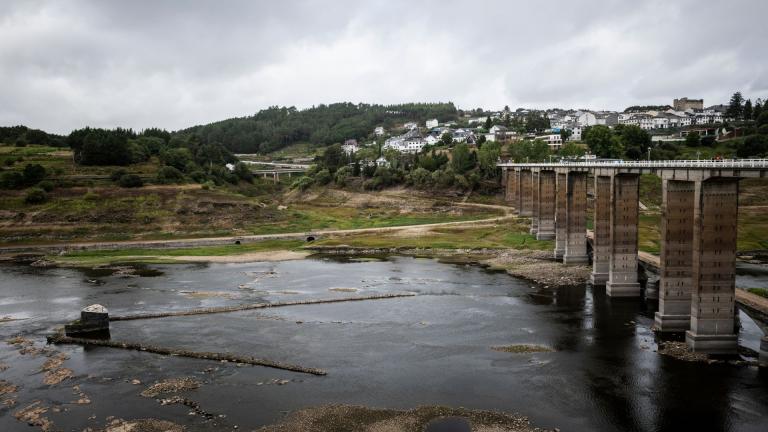 A partially dry riverbed with exposed rocks and a tall concrete bridge, with a hillside town and green trees in the background under a cloudy sky.
