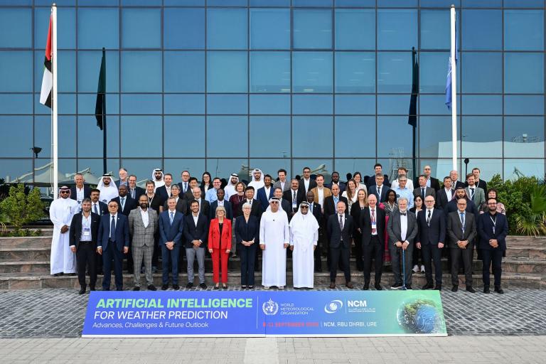 A large group of people in formal attire pose for a photo outside a modern glass building at an AI for weather prediction conference, with event banners and flags visible.