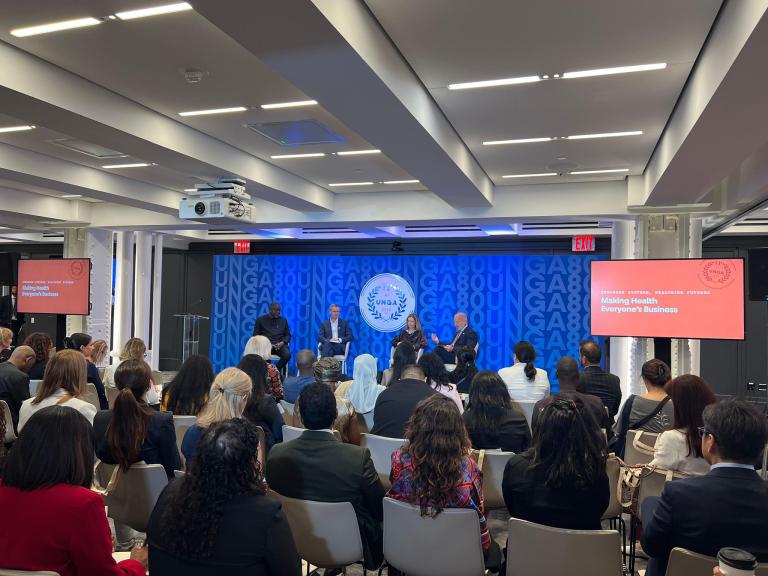 A diverse audience sits in a conference room listening to a panel discussion titled "Making Health Everyone's Business" at a UNGA event.