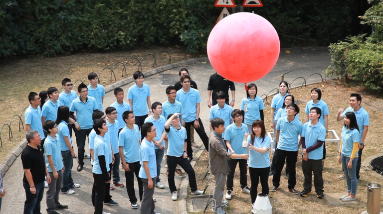 A group of people in matching blue shirts gather outdoors around a large red balloon, with some looking up and others facing the balloon.