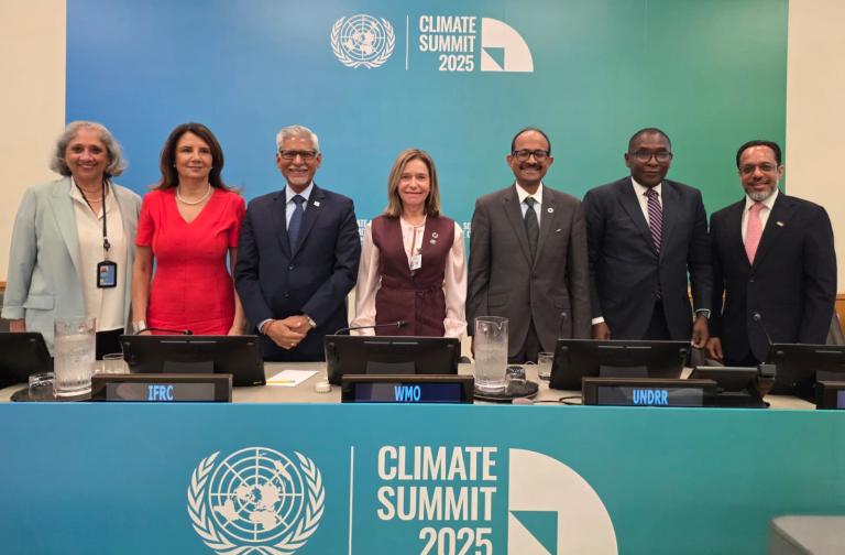 Seven people in business attire stand behind a desk at the Climate Summit 2025, with United Nations and summit logos visible on the blue-green backdrop.