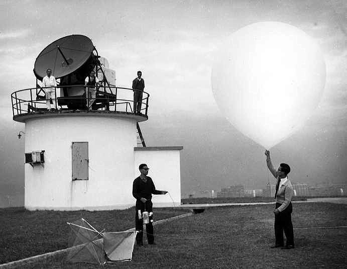Four people work at a weather station; one person holds a large weather balloon, while others stand near an observation tower with a radar dome.