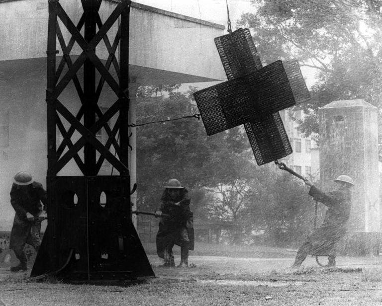 Three people in protective gear operate a large cross-shaped device attached to a structure in rainy or misty conditions, with water spraying around them.