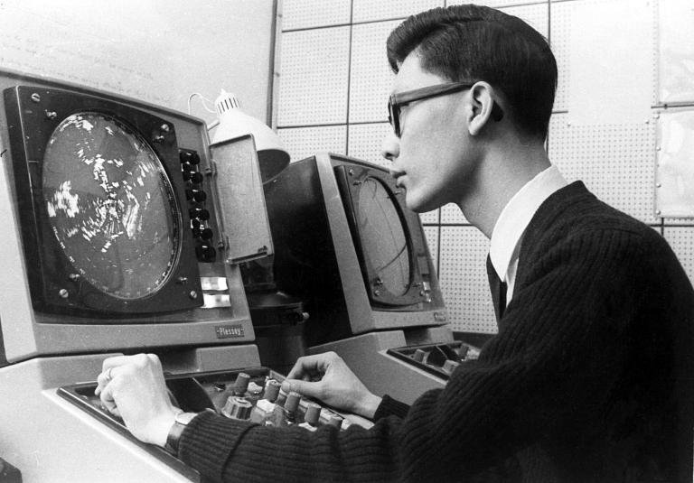 A man wearing glasses operates controls at a radar or computer console with a large circular screen displaying data.