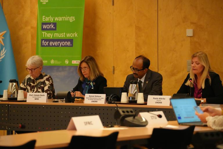 A panel of four speakers sits at a conference table during an event on early warning systems. Behind them is a green banner reading “Early warnings work. They must work for everyone.” The panelists are seated in front of nameplates showing their affiliations with WMO and UNDRR, and microphones and laptops are visible on the table.