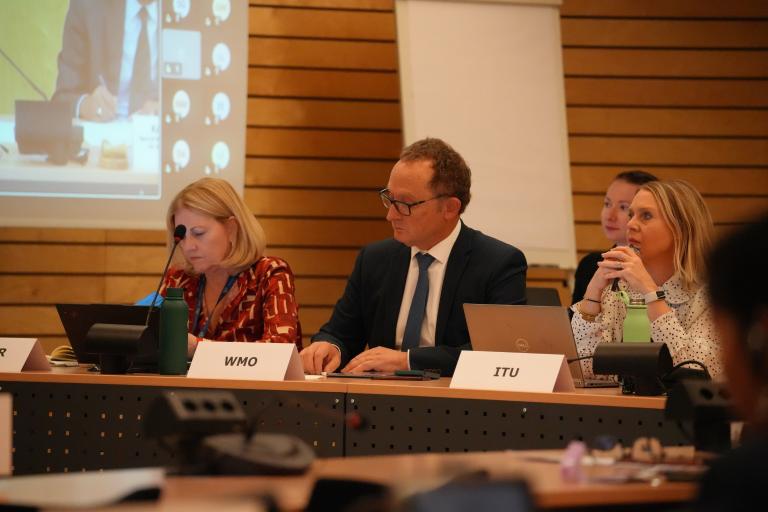 Three panelists sit at a conference table during a formal meeting, each with laptops and microphones. Nameplates in front of them read “UNDRR,” “WMO,” and “ITU.” A presentation is projected on a screen in the background, and other participants are partially visible in the room.