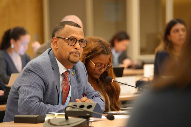 A man in a suit and glasses speaks into a microphone during a formal meeting, with another participant beside him taking notes. Several other attendees are visible in the background, seated and using laptops in a conference room setting.