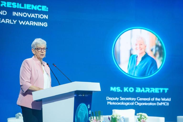 A woman speaks at a podium onstage, with a large screen behind her displaying her name, title, and photo: "Ms. Ko Barrett, Deputy Secretary General of the World Meteorological Organization.