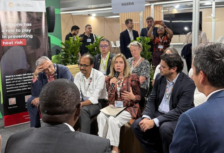 A group of people sits in a circle having a discussion at an event; a sign reading "NIGERIA" and a poster about heat risk are visible in the background.