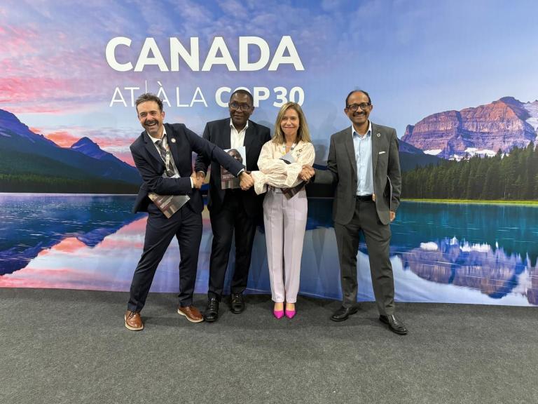 Four people in business attire stand in front of a "Canada at/la COP30" backdrop, posing together and holding a large circular object.