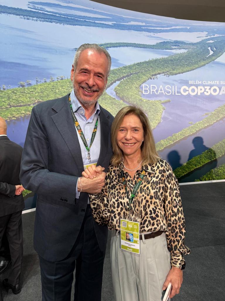 A man and a woman stand together, shaking hands and smiling at the Belém Climate Summit, with a Brasil COP30 sign and river scenery in the background.