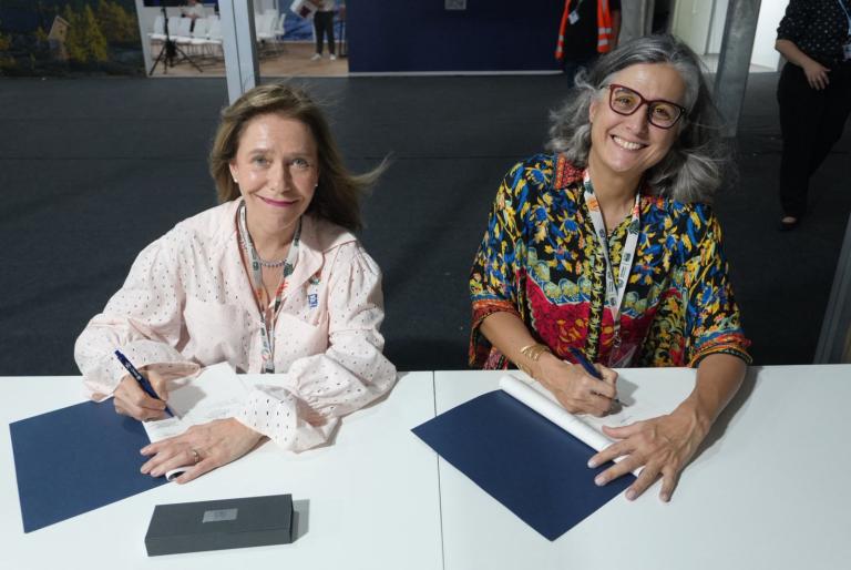 Two women sit at a table, each holding a pen and signing documents, facing the camera and smiling at an indoor event.