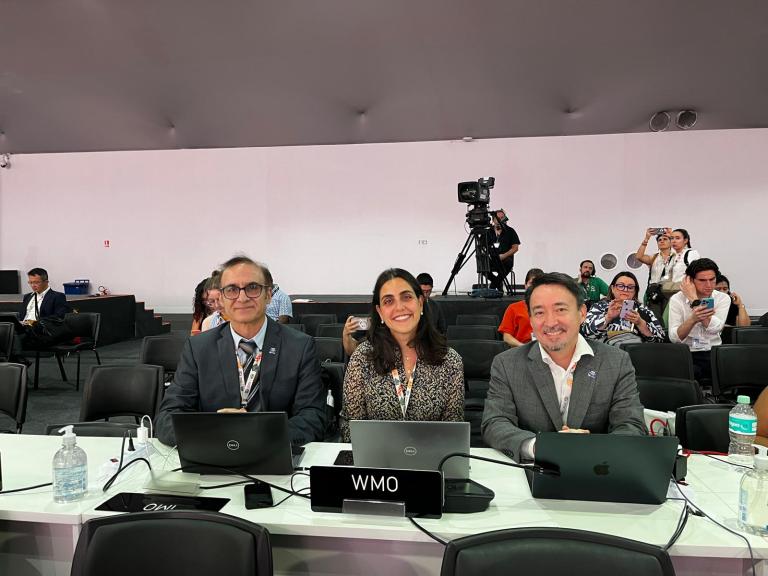 Three people sit at a table labeled "WMO" with laptops in front of them at a conference; other attendees and a camera crew are visible in the background.