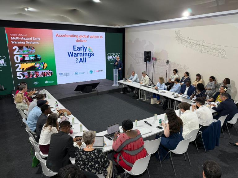 A group of people sit around a U-shaped table at a conference, facing a presentation screen displaying "Early Warnings for All" and related climate action messages.