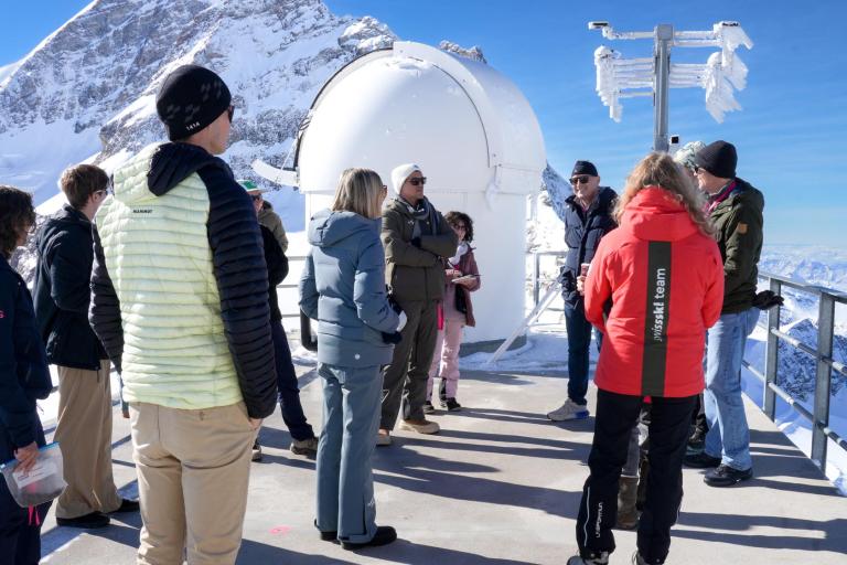 A group of people in winter clothing stand on a snowy mountain observation deck near a white dome structure, listening to a guide.