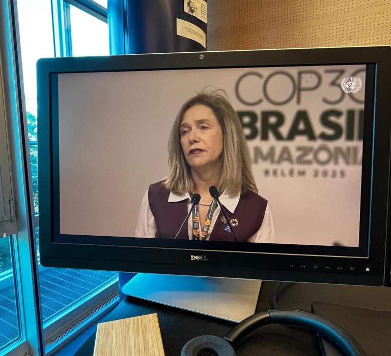 A woman speaks at a podium during COP 30, with a sign behind her reading "COP 30 Brasil Amazônia Belém 2025," displayed on a computer monitor.