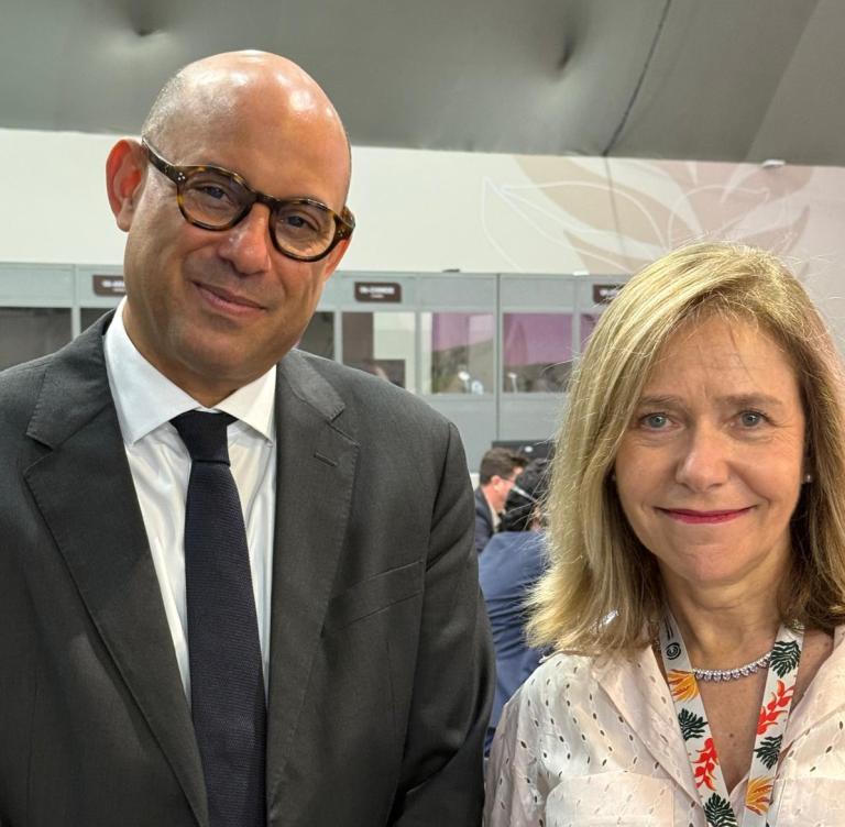 A man in a suit and glasses stands next to a woman in a light blouse at an indoor professional event.