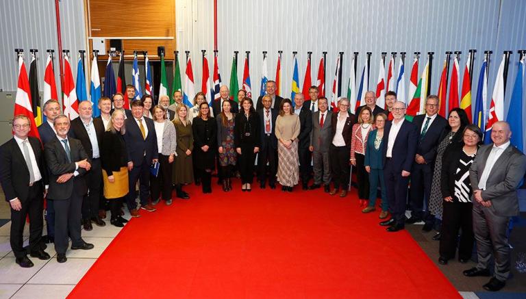 A large group of people in formal attire stands on a red carpet in front of a display of various international flags.