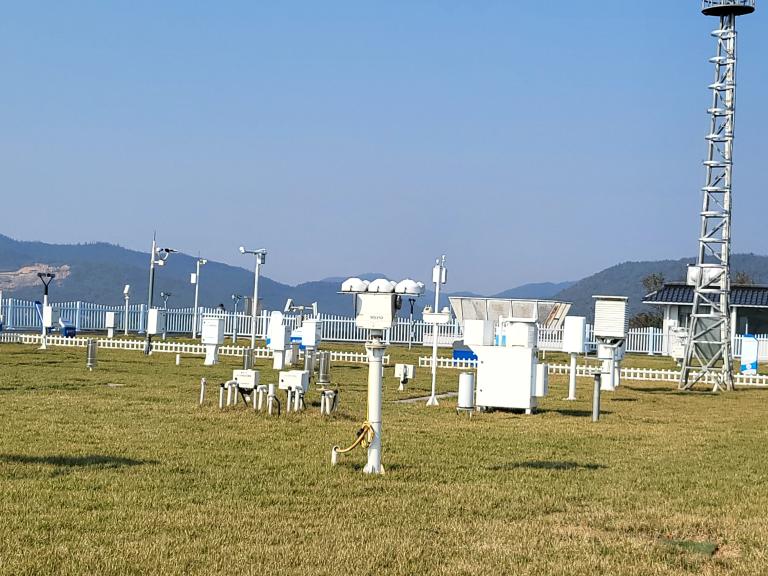 A weather station with various meteorological instruments and sensors arranged on a grassy field, surrounded by a white fence and hills in the background.