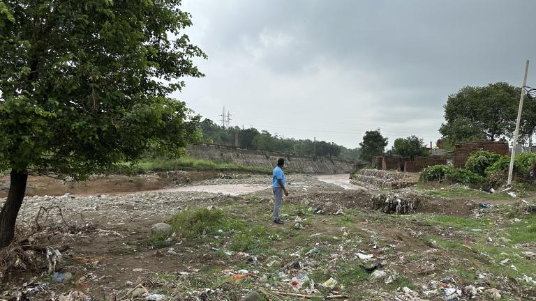 A person in a blue shirt stands near a litter-strewn riverbank, with trees, scattered trash, and cloudy skies in the background.