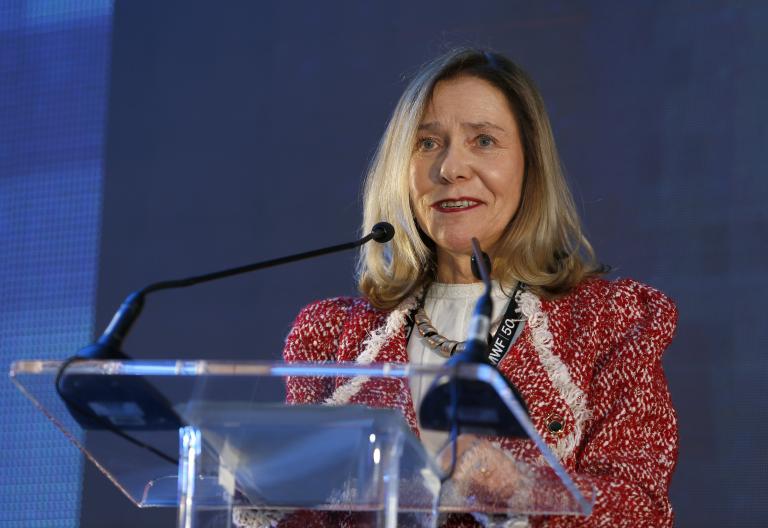 A woman in a red and white jacket speaks at a clear podium with two microphones in front of a blue and gray background.