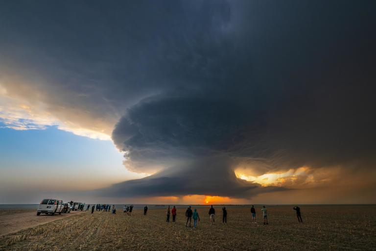 A group of people stands on open ground, watching a large, dramatic supercell thunderstorm cloud form during sunset.