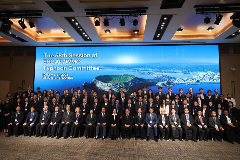 A large group of people in formal attire pose for a group photo in front of a screen displaying "The 58th Session of ESCAP/WMO Typhoon Committee, Jeju Island, Korea.