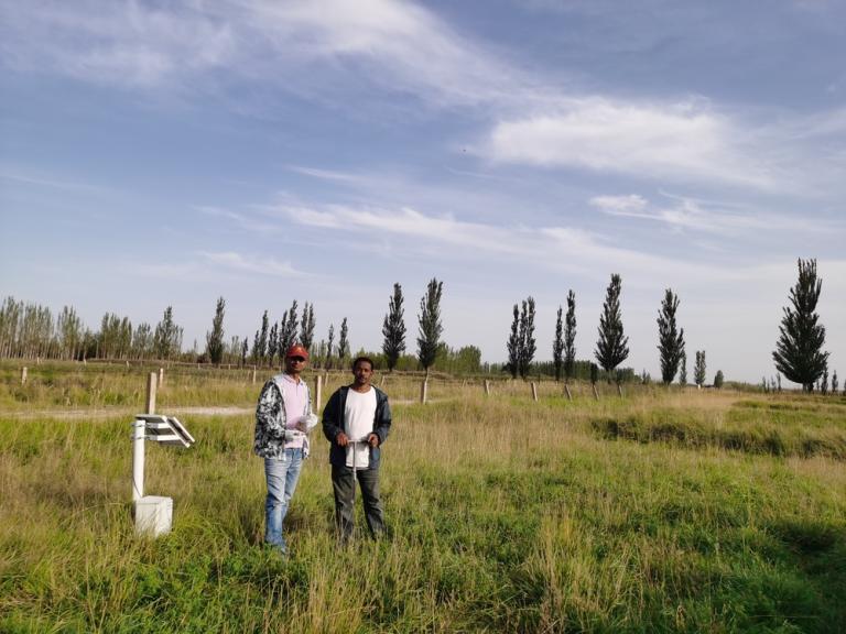 Two people stand in a grassy field near a metal weather instrument, with trees and a blue sky with scattered clouds in the background.