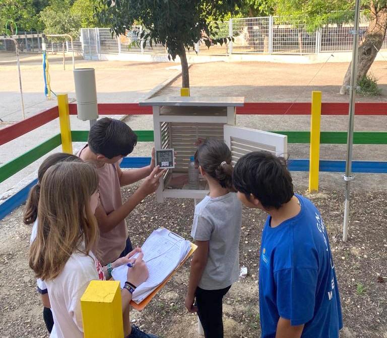 Four children observe and record data from a weather station instrument outdoors, with one holding a device and another writing on a clipboard.