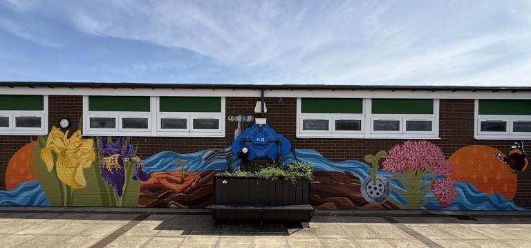 A mural with colorful flowers, plants, and a butterfly is painted on a brick wall with small windows, featuring a large blue pipe in the center above a planter box.