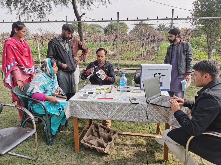 A group of people gather around a table outdoors, examining equipment and using a laptop, with demonstration materials and a poster on the table.