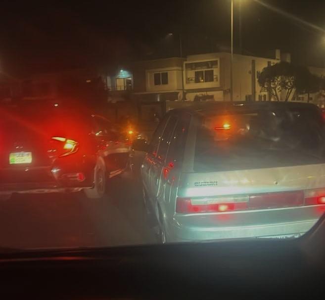 Nighttime traffic scene showing several cars with brake lights illuminated, viewed from inside a vehicle, with buildings and streetlights visible in the background.