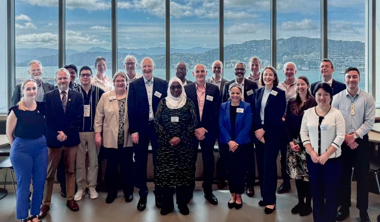 A diverse group of people in business attire pose for a group photo indoors, with large windows showing hills and a cloudy sky in the background.